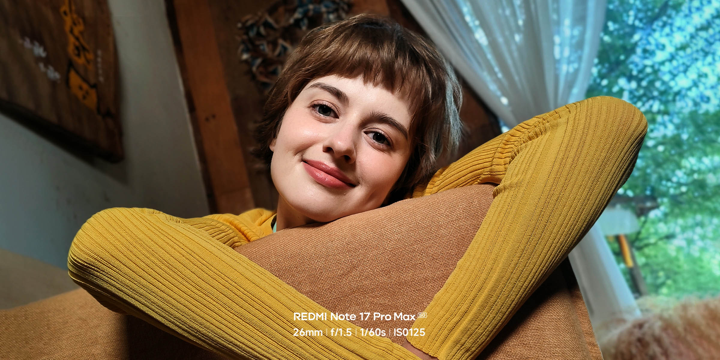 A young woman smiles while leaning on the sofa, wearing a yellow sweater, with green trees outside the window and soft indoor lighting in the background.