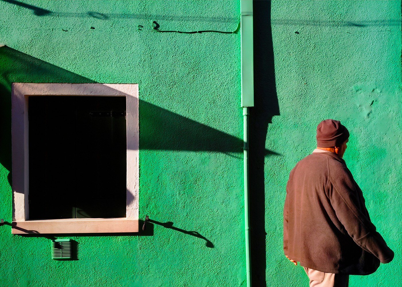 一個穿著深色外套和帽子的男性站在綠色牆壁旁,背景中有一個窗戶和一根綠色管道,陽光投射出陰影。