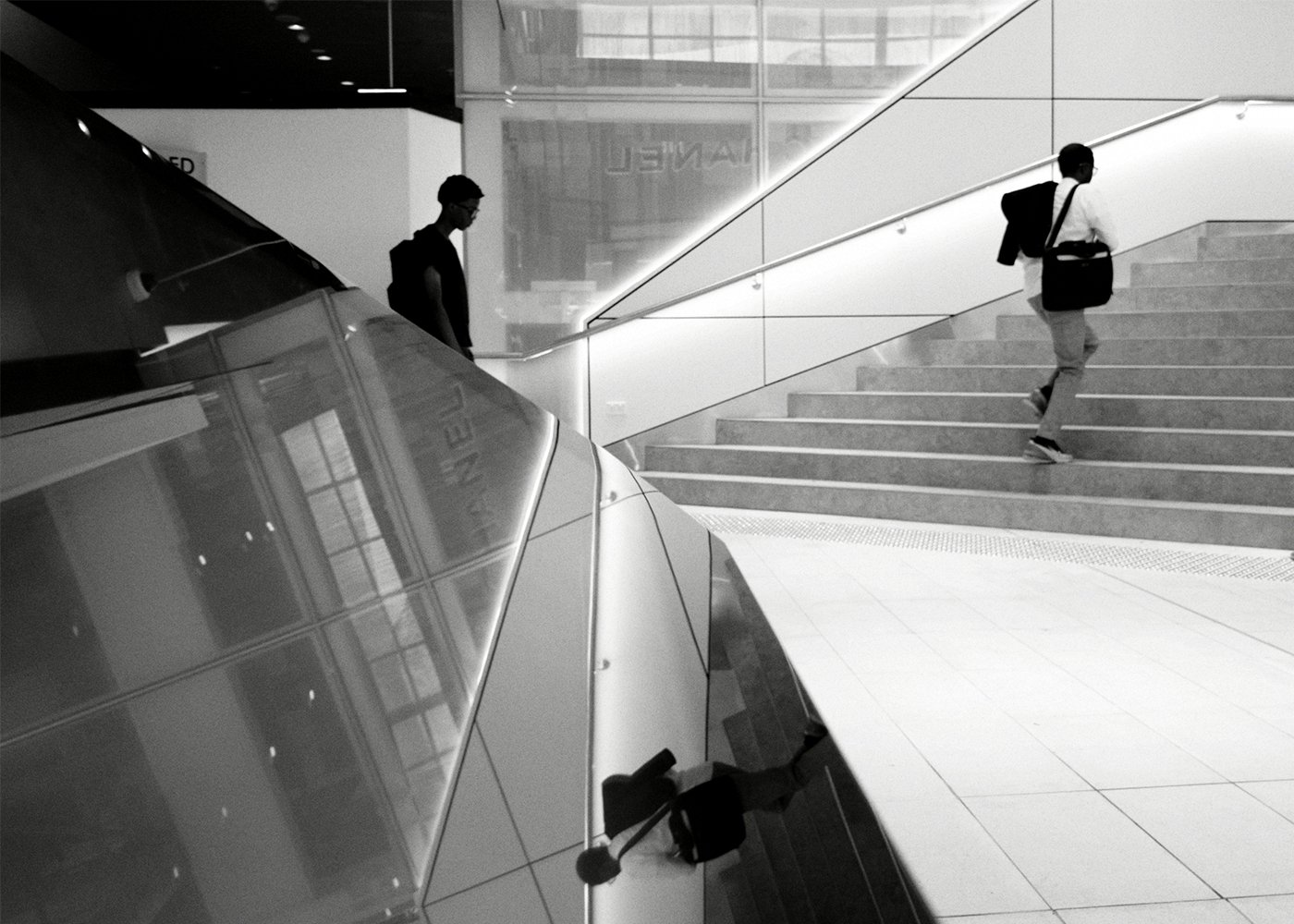 兩個人在現代建築的階梯上行走,左側有一個人正走下階梯,右側有一個人正向上走。背景是光滑的玻璃牆面,反射出周圍環境。整體畫面呈現黑白色調,營造出簡約而現代的氛圍。