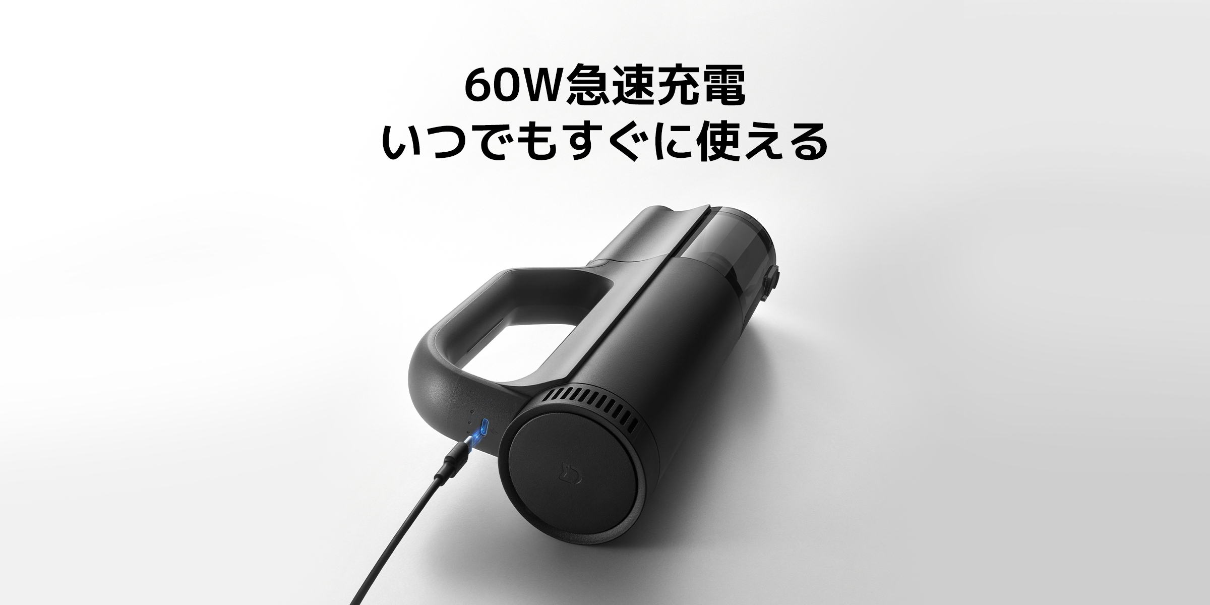 これは黒いハンドヘルド掃除機で、充電中です。掃除機のデザインはシンプルで、充電ポートの横に青いインジケーターライトがあります。背景はシンプルな白です。