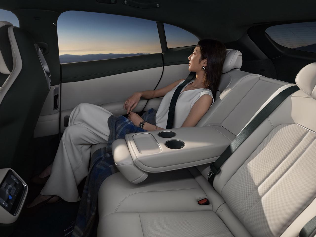 A woman is sitting in the back seat of a car, wearing a white top and light-colored pants, with two cup holders beside her, and in the distance, mountains can be seen outside the window.