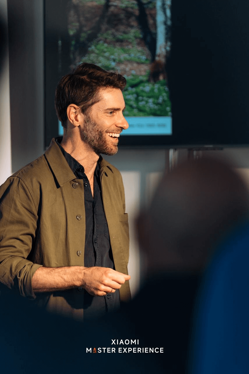 A man wearing a green jacket is giving a speech, with images related to nature displayed in the background, and the text "XIAOMI MASTER EXPERIENCE" appearing at the bottom of the screen.