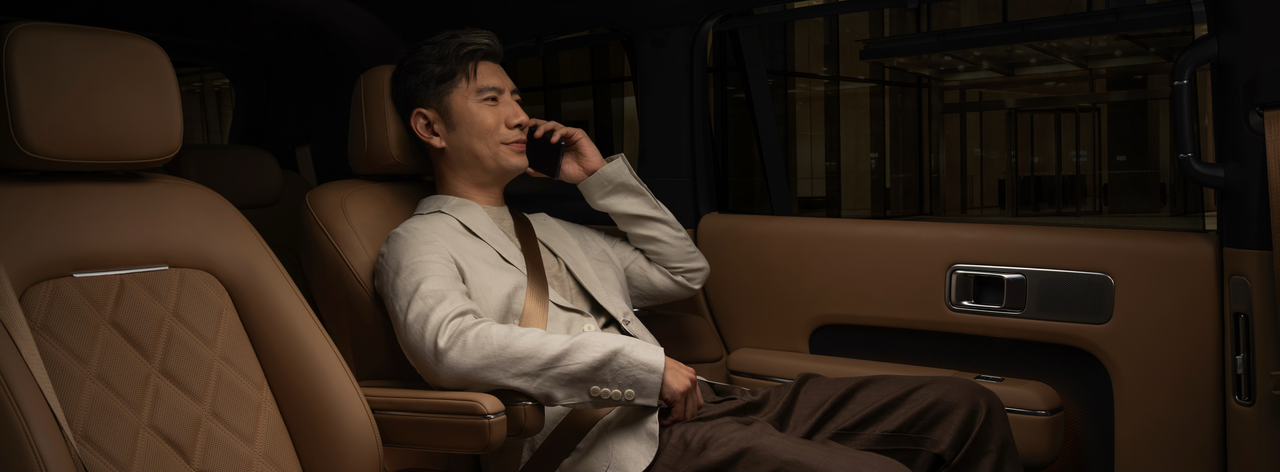 A man is sitting in the back seat of a luxury car, smiling while on the phone, with warm brown tones in the surrounding interior. The environment outside the car window is blurred, creating a cozy atmosphere.
