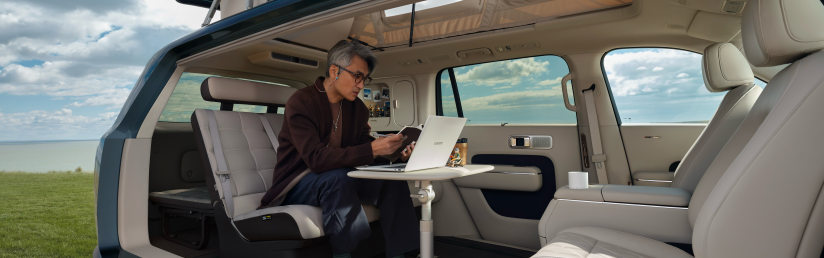 A man is sitting inside a car, using a laptop, with a small table nearby. Outside the car window, a blue sky and a sea view can be seen, and the interior of the car is decorated in a modern style with light-colored leather seats.