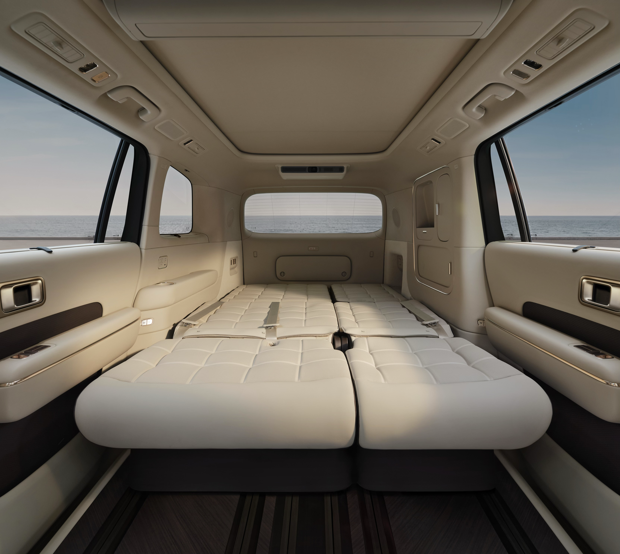 The interior space showcases the back seat of a car, with the seats folded down to create a flat resting area. The decor features beige leather, and through the windows, a view of the ocean can be seen in the background.