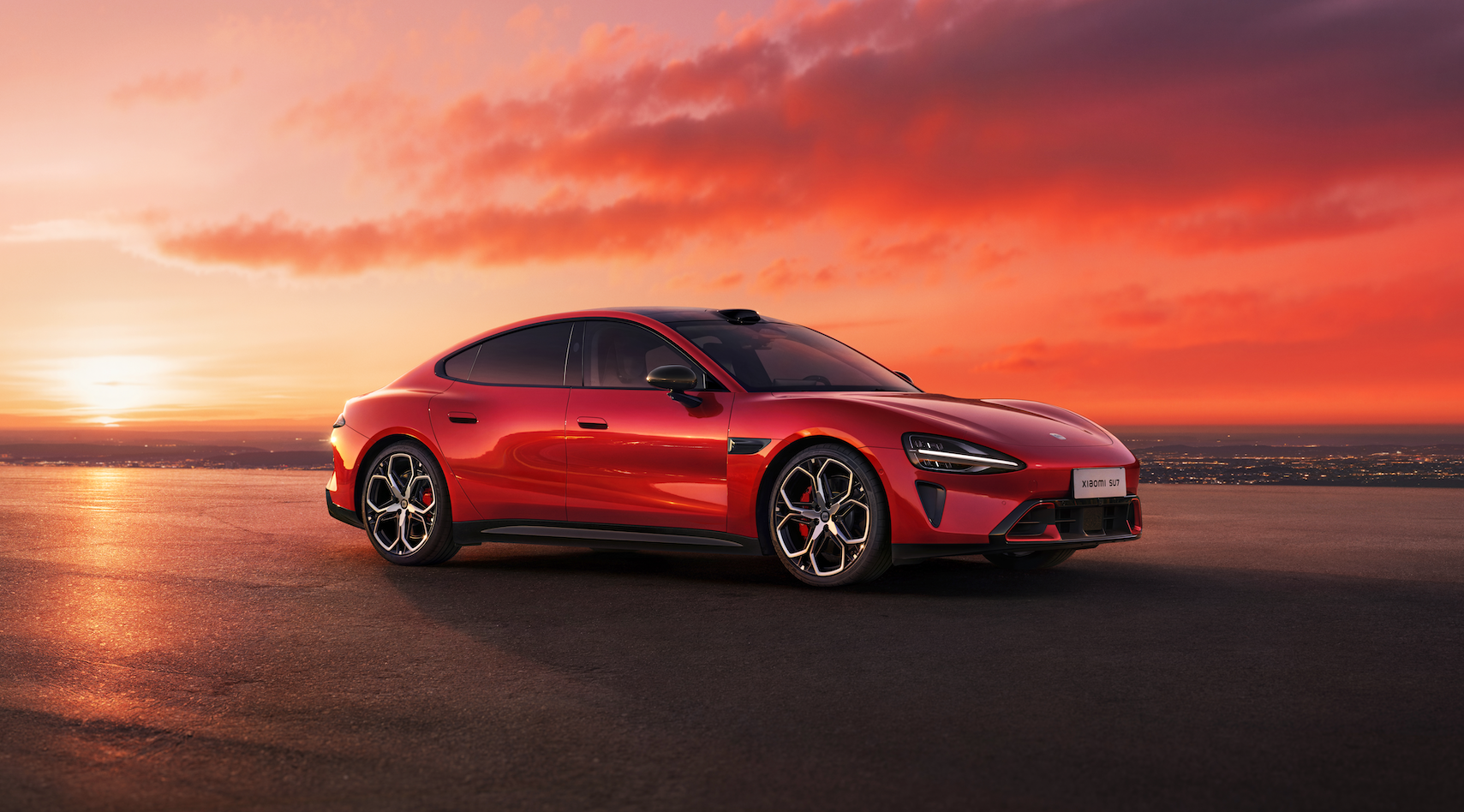 A red sports car is parked on a flat surface, with a stunning sunset in the background, orange and purple clouds in the sky, and the city lights visible in the distance.