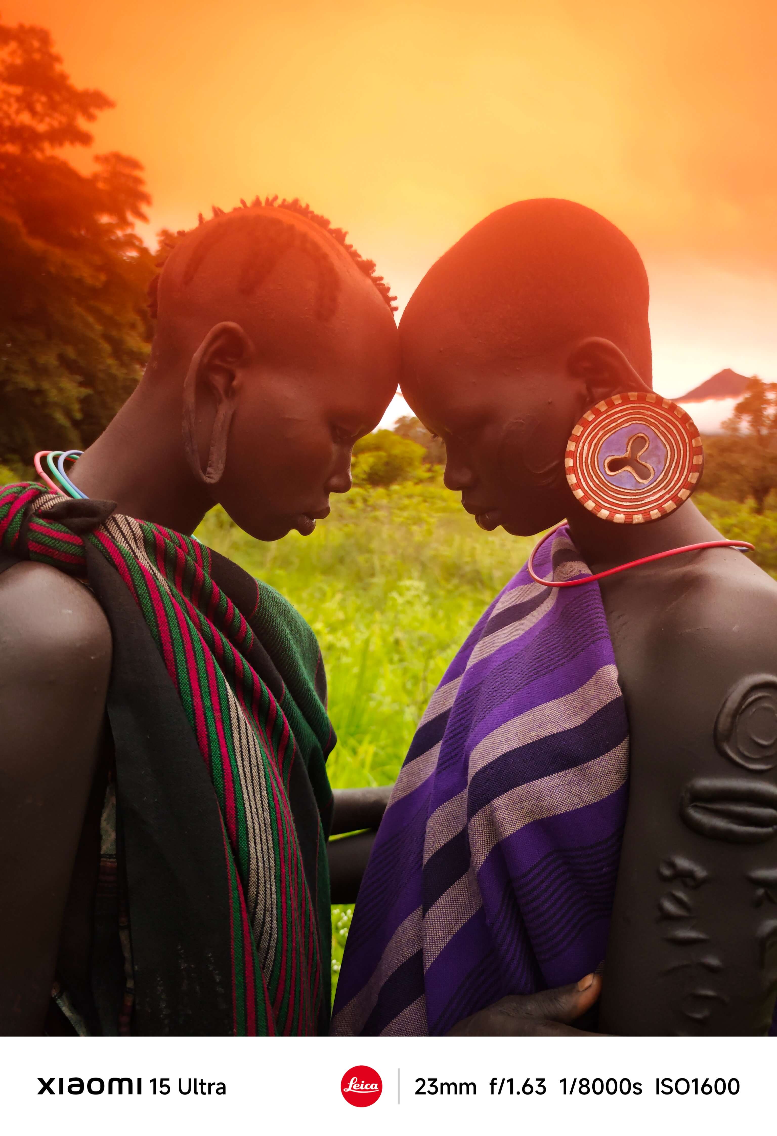 Two people dressed in traditional attire stand together, with a green meadow and an orange sky at sunset behind them. Their bodies are adorned with decorative patterns and earrings, showcasing cultural characteristics.