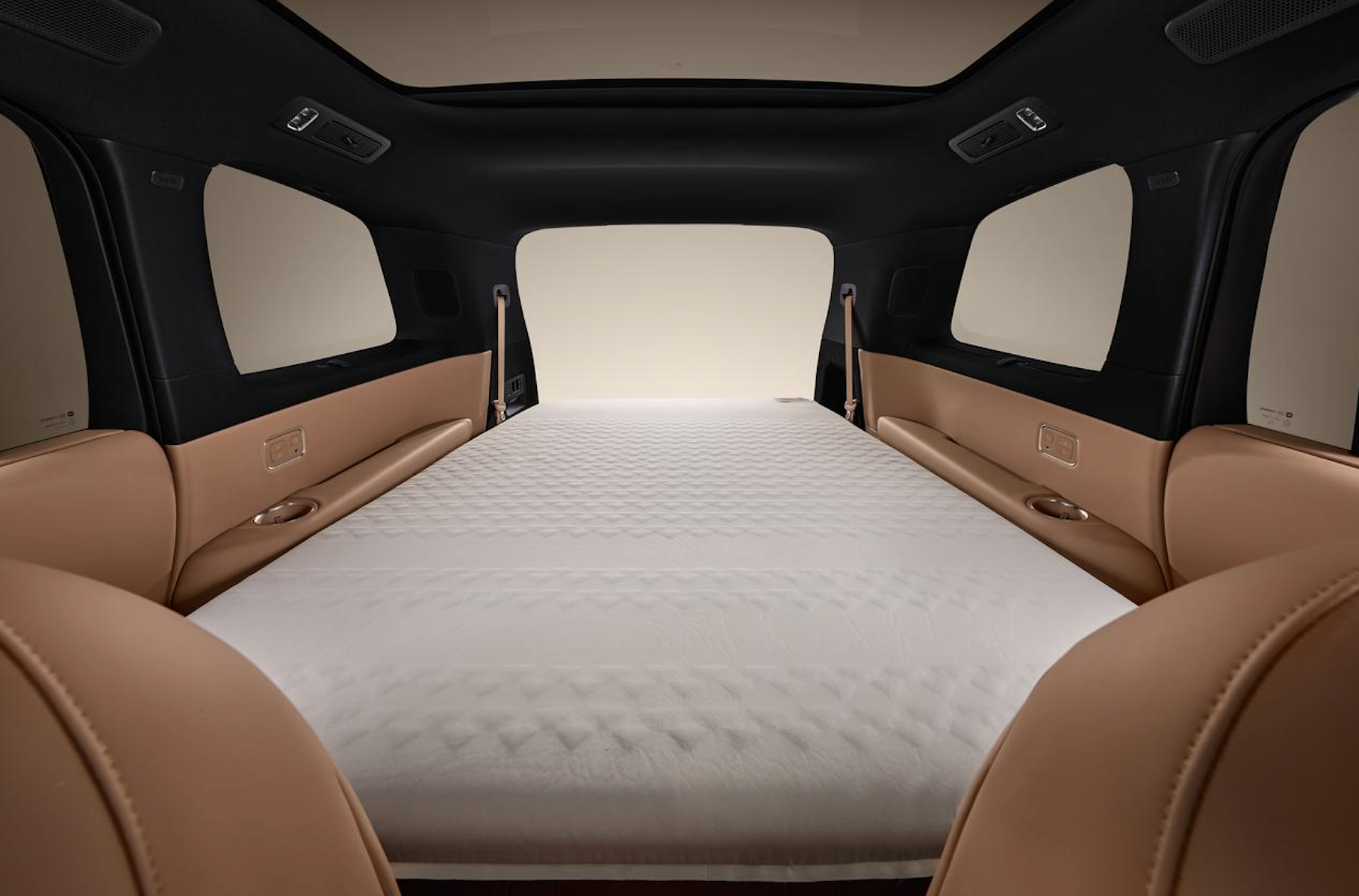 The interior of the car is arranged to look like a bed, with a white mattress placed at the back of the car, surrounded by brown seats and interior decor. The windows are large, allowing for bright light.