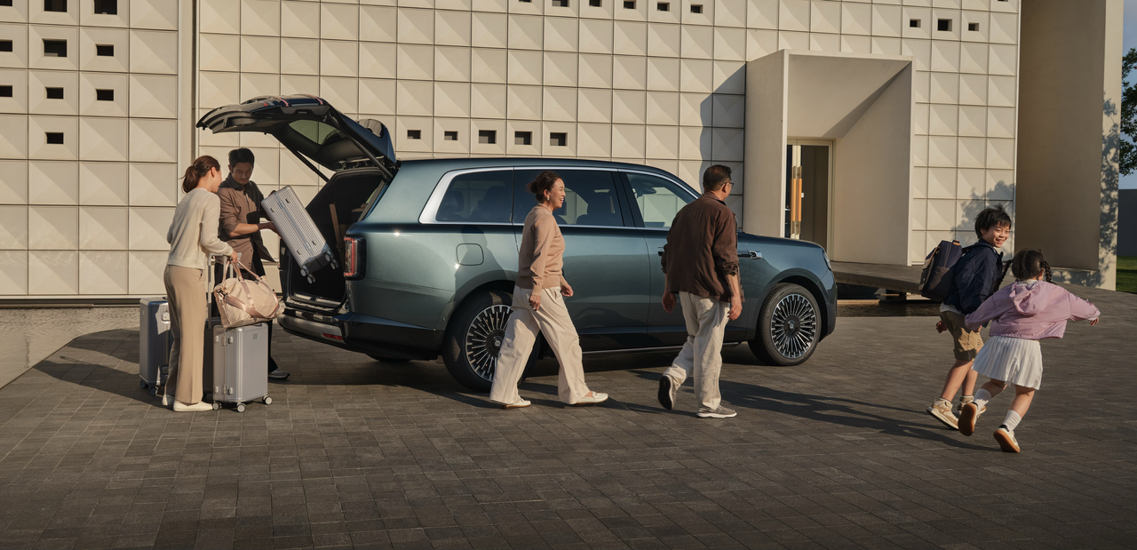 A family is busy loading luggage next to a blue SUV, with a woman putting bags into the trunk while another man assists her. Two children are playing nearby, with modern buildings and a paved stone road in the background.