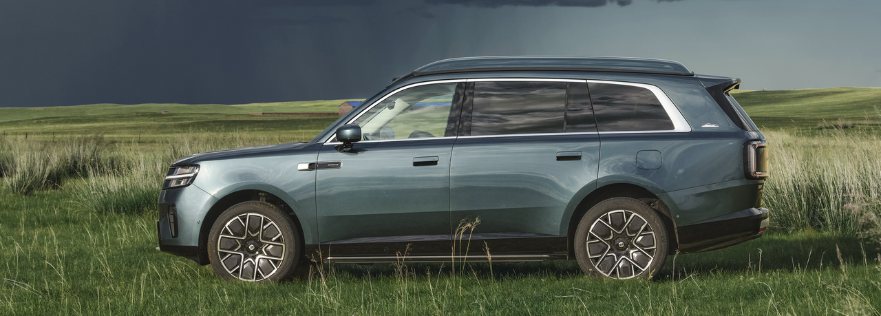 A blue SUV is parked on a vast lawn, with a backdrop of a cloudy sky and tall grass surrounding it.