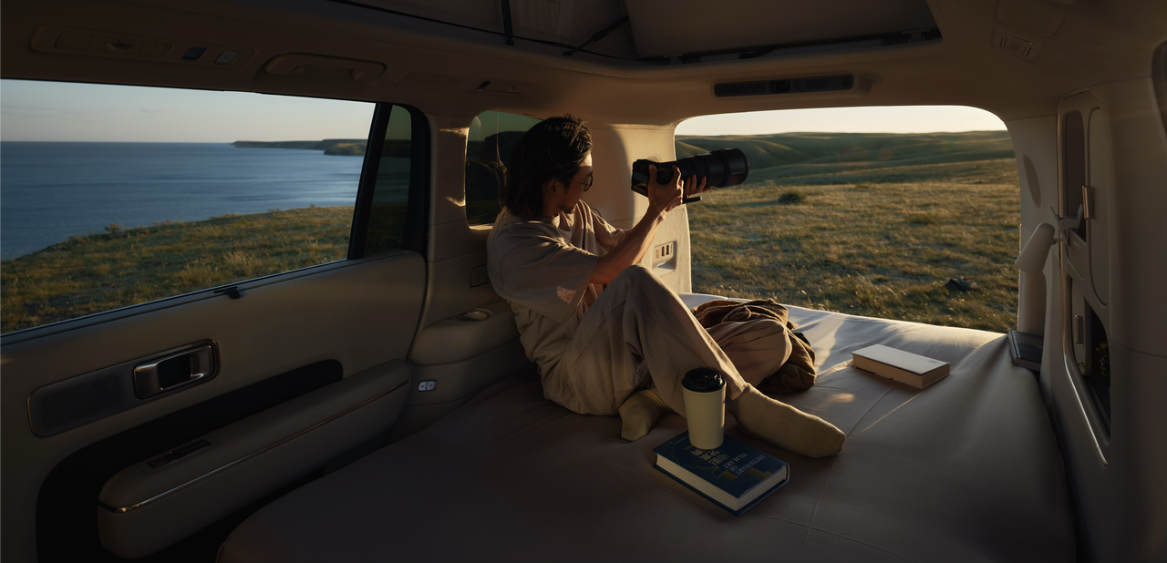 A person is sitting inside a car, holding a camera, preparing to photograph the scenery outside the window. Outside the car window is the ocean and grasslands, with sunlight pouring into the car, creating a warm atmosphere. Inside the car, there is a book and a coffee cup.