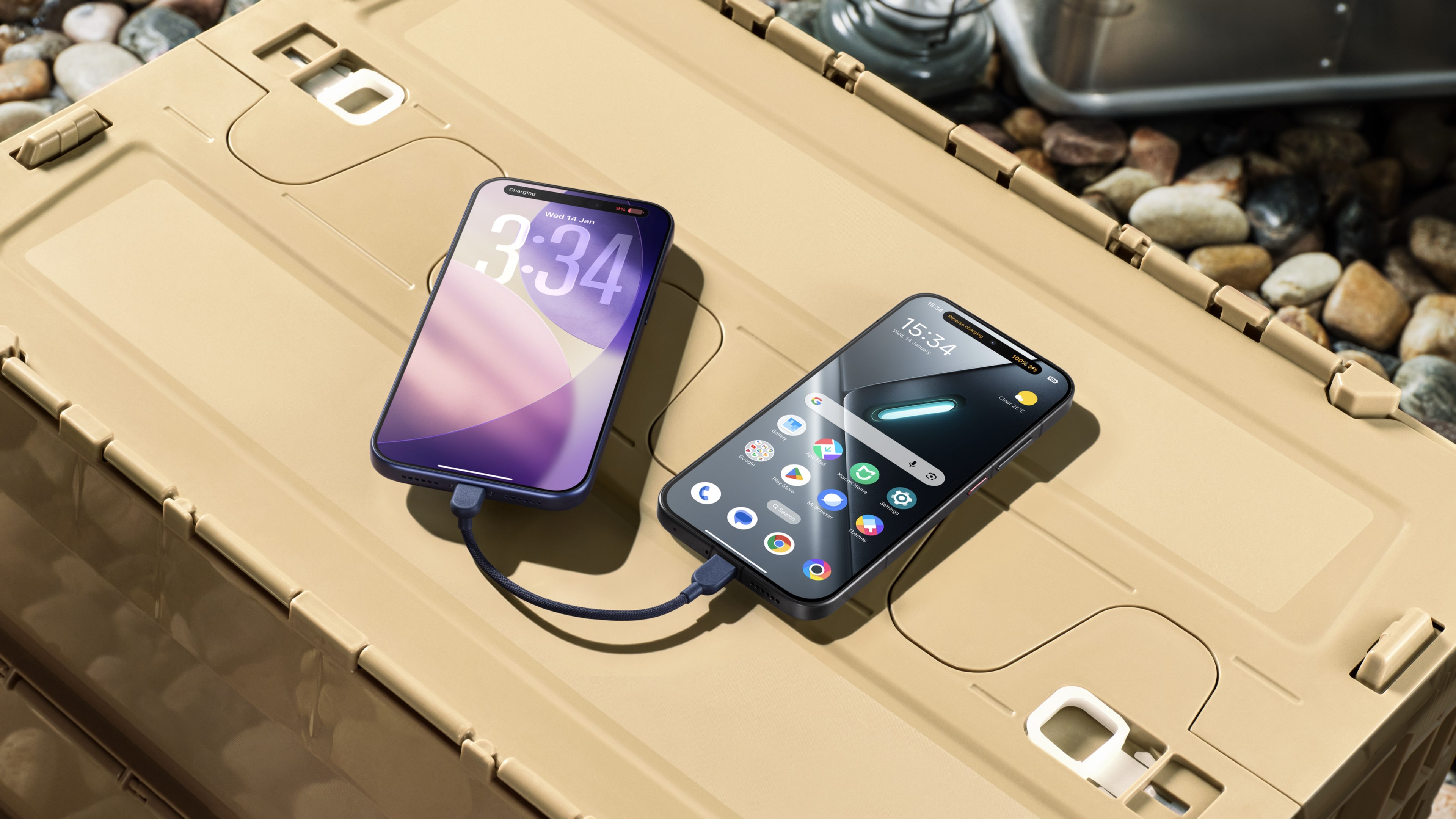 Two smartphones are placed on a brown box, connected together by a data cable. One smartphone's screen shows the time as 3:34, while the other displays the time as 15:34, with a background of some small stones.