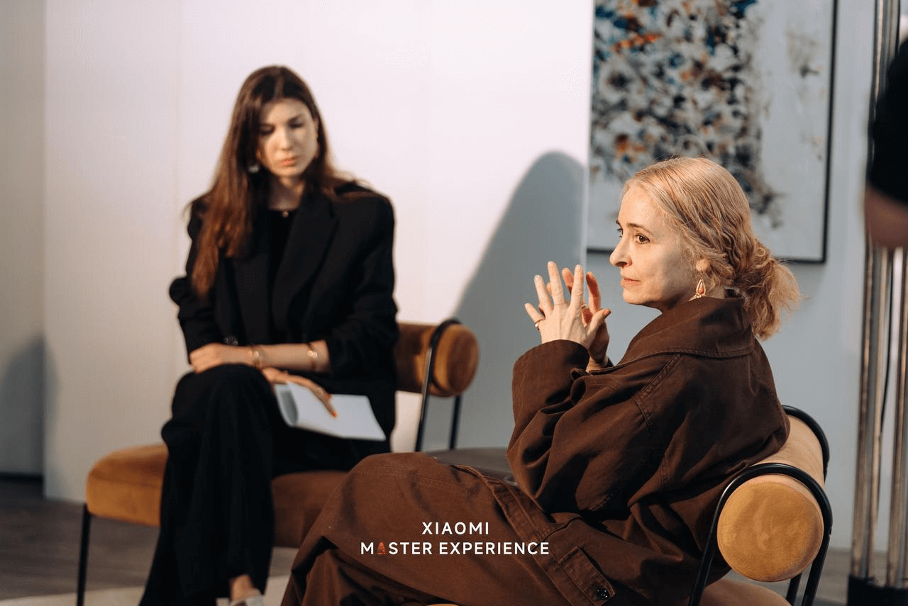 Two women are sitting on chairs, one wearing a black suit and the other wearing a brown coat, engaged in conversation, with modern art pieces in the background.
