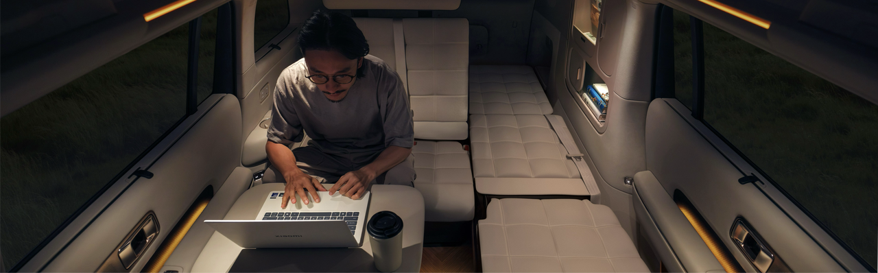 A man is sitting on the sofa inside the car, focused on working on his laptop, with a coffee cup placed beside him. The interior of the car is simple and comfortable, with soft lighting.