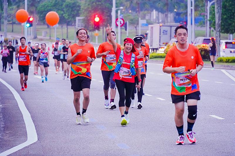 A group of people wearing orange running suits is participating in a marathon on the road, with traffic lights and some passing vehicles in the background.