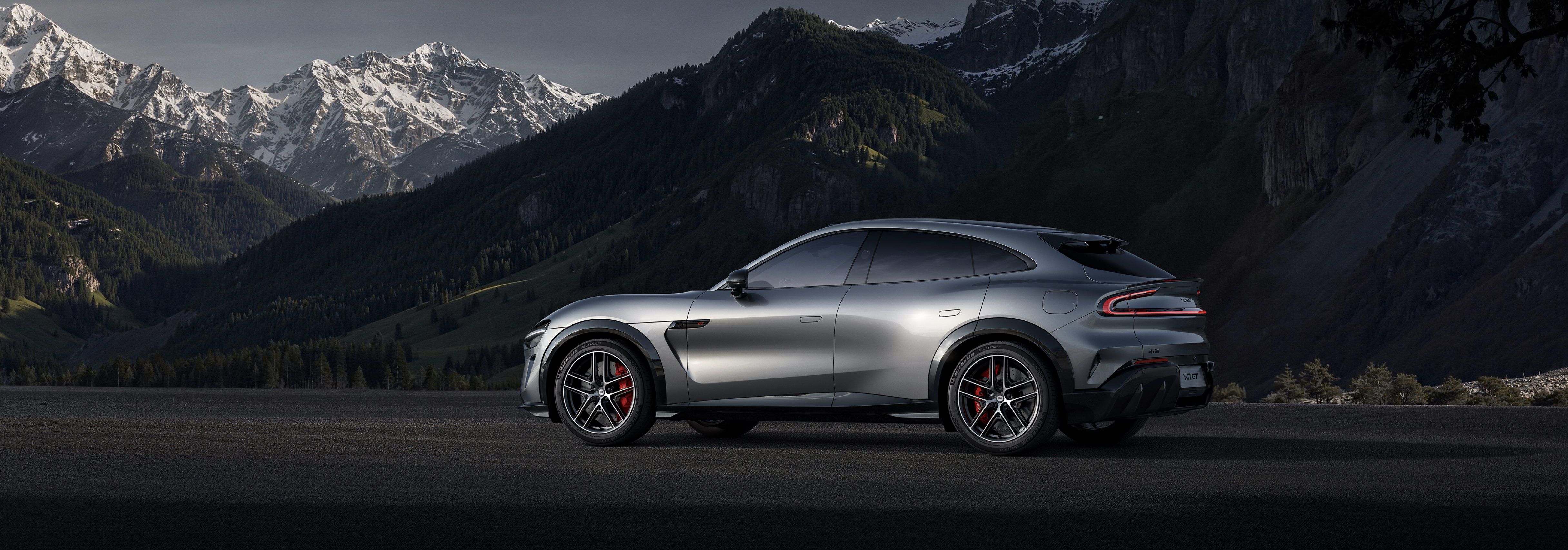 A silver sports car is parked on the mountain road, with towering snow-capped mountains and dense forests in the background.