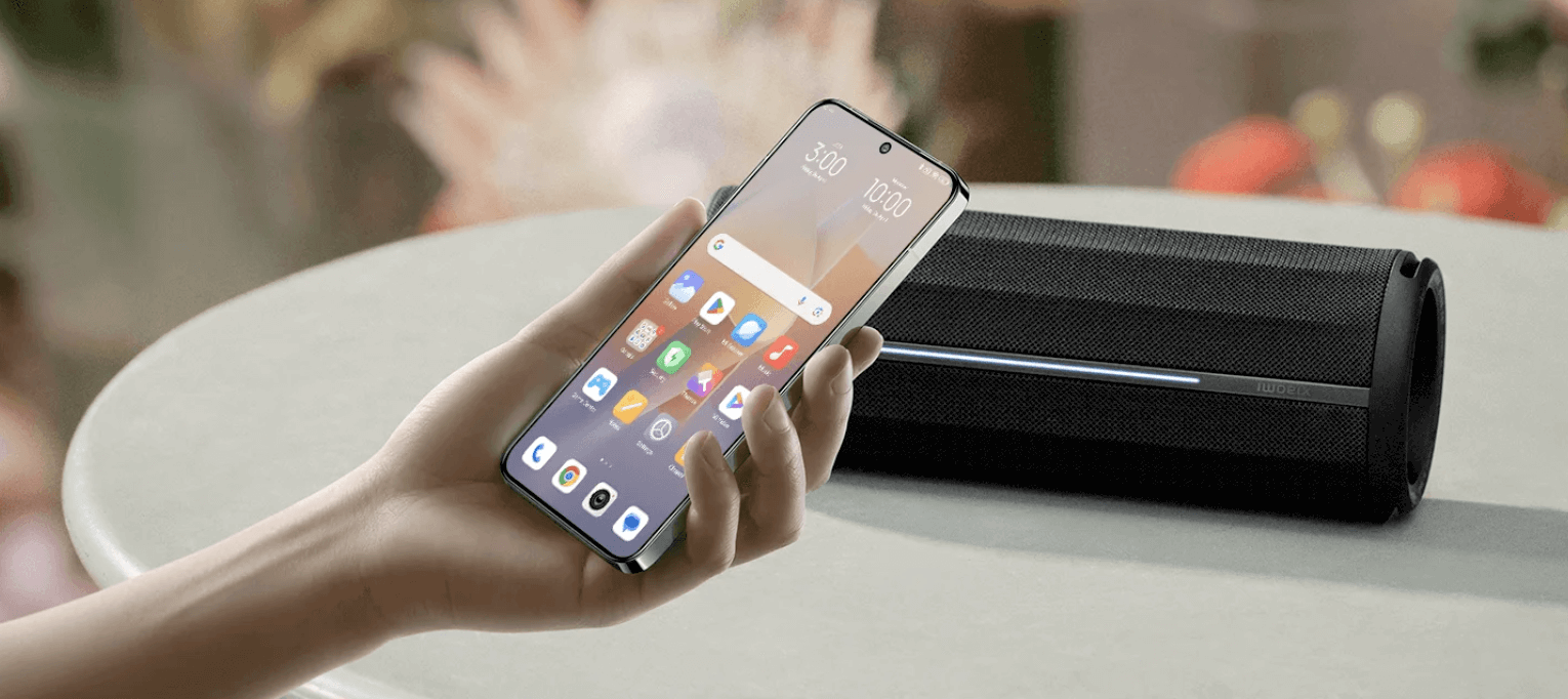 A hand is holding a smartphone, the phone's screen displays multiple app icons, next to it is a black Bluetooth speaker, and the background is blurred.