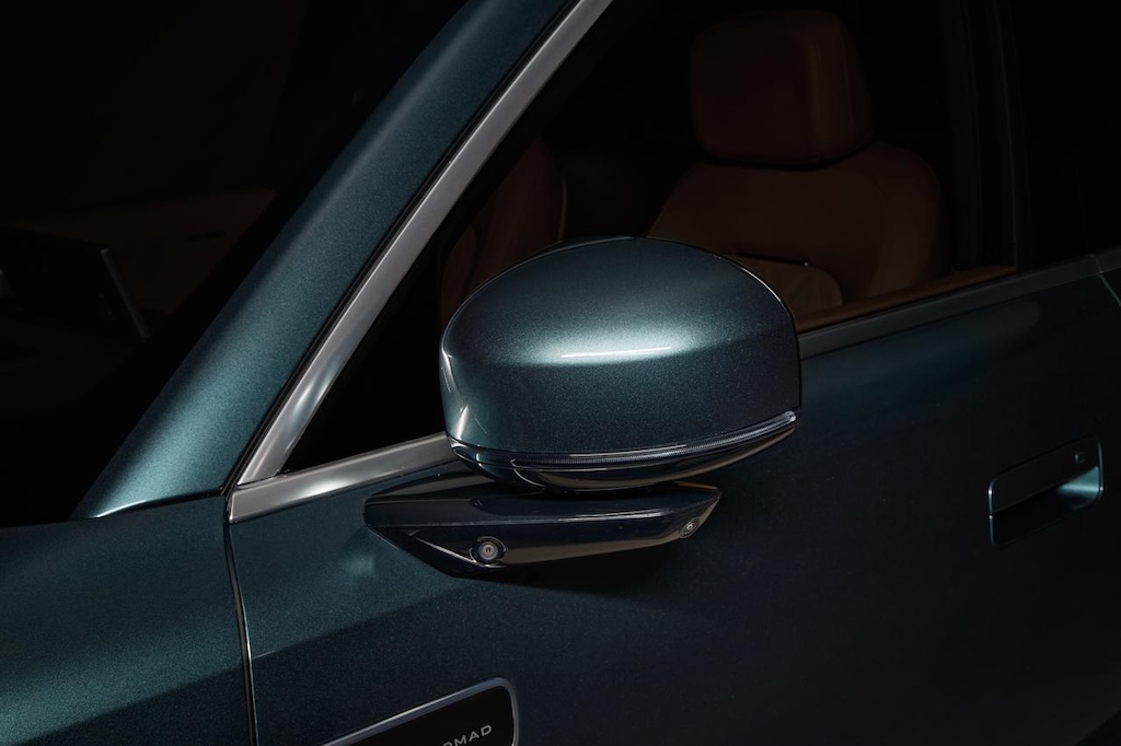 A close-up of the car's side mirror, with the mirror and window frame displaying a dark metallic sheen, set against a black background, and brown seats visible inside the car.