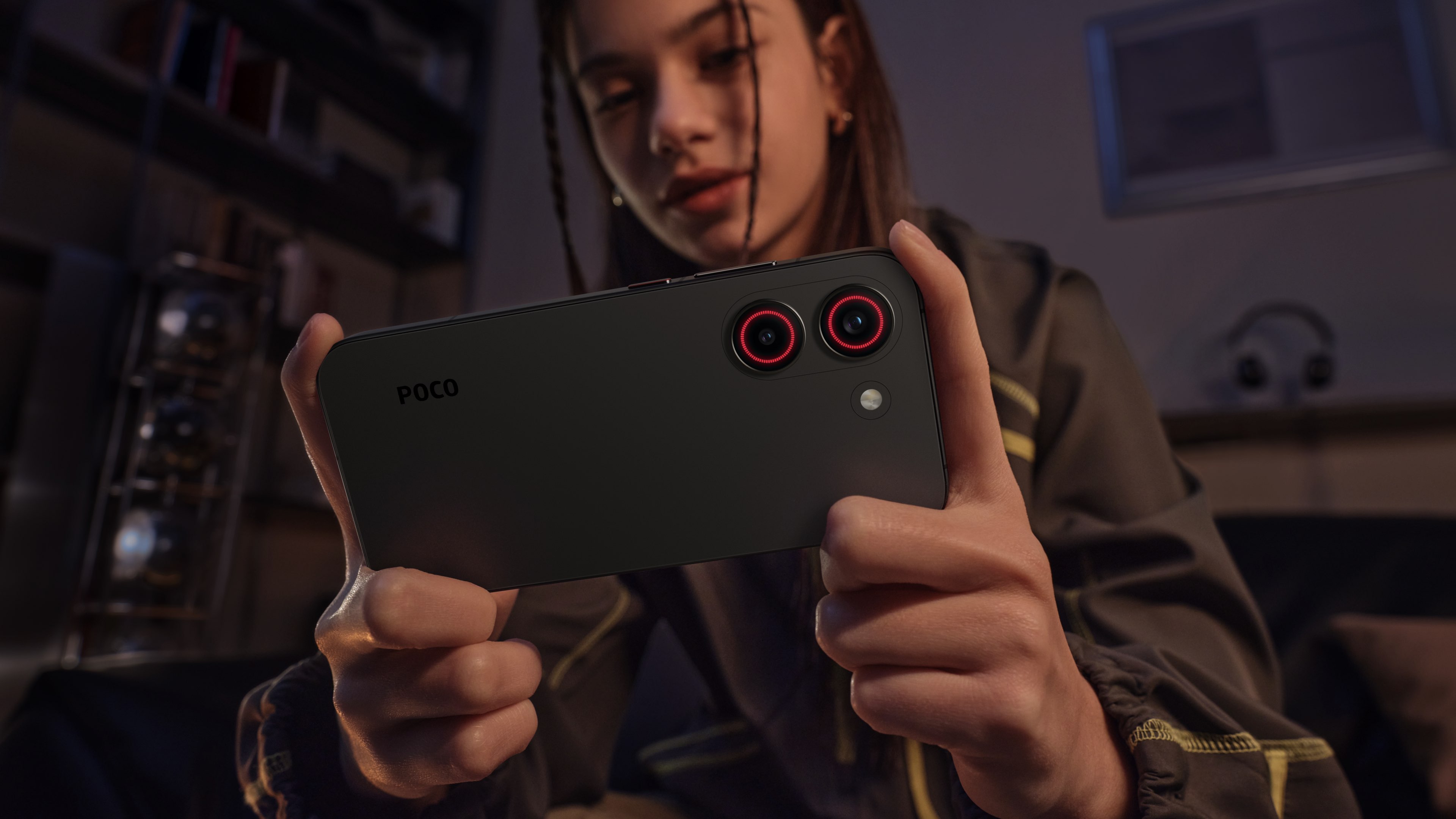 A young woman is holding a black phone in a low-light environment, with two red lenses on the back of the phone, her expression focused. In the background, some blurred home elements and headphones can be seen.