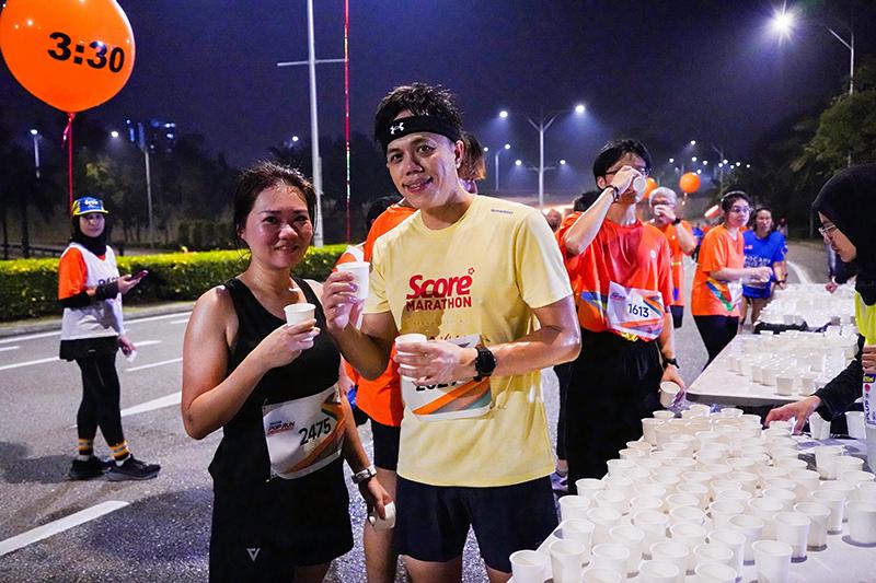 The athlete stops during the nighttime marathon to grab a drink, surrounded by many cups and other competitors, with a brightly lit street in the background and a balloon marked "3:30."