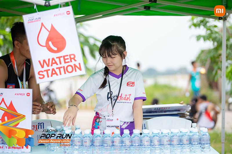 Volunteers are preparing to distribute bottled water next to the water station, with a background of green trees and a crowd of people participating in the event.