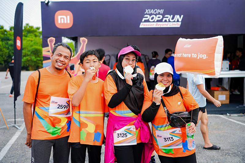 A group of participants dressed in orange sportswear posed for a photo during the "XIAOMI POP RUN" event, with the event banner and other participants in the background. Some held up a sign that read "I'm not here to jog."