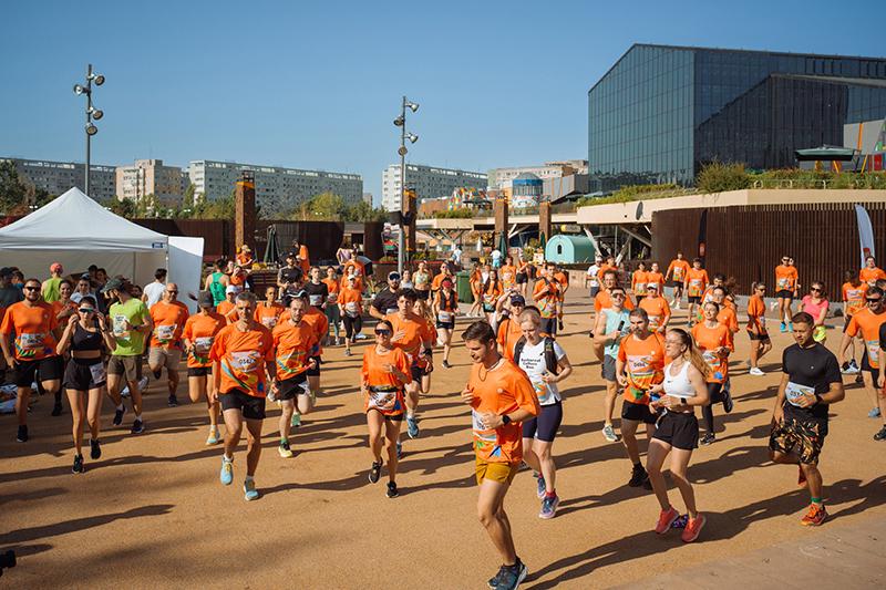 Many participants wearing orange T-shirts are running outdoors, with the city buildings and blue sky in the background, and there are tents and other participants on the field.
