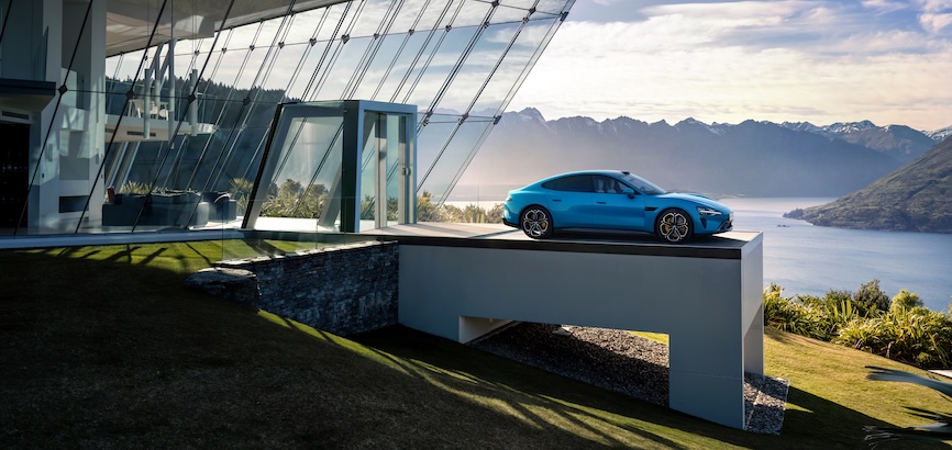 A blue sports car is parked on the terrace of a modern building, with a backdrop of lakes and distant mountains. The building features extensive glass design, and the surrounding environment is lush and green.