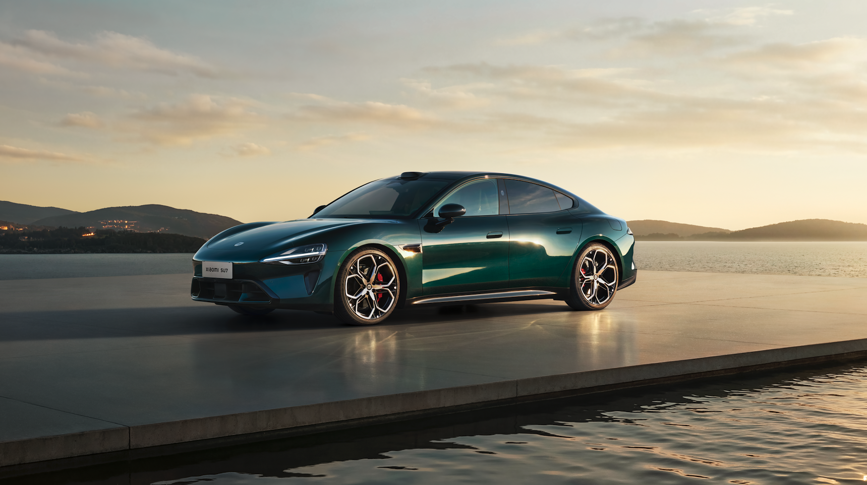 A green sports car is parked on a modern platform by the water, with a tranquil lake surface and distant hills in the background, as the sunset casts a soft glow.