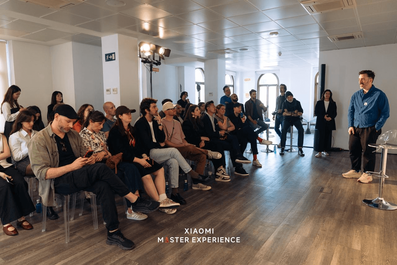 A group of people is attending a meeting, listening to a speaker in a blue coat, with a bright indoor environment in the background. The floor is wooden, and there is a sign on the wall that says XIAOMI MASTER EXPERIENCE.