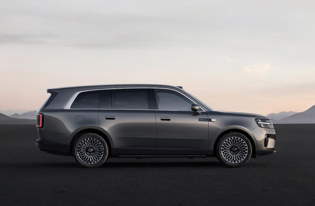 A gray SUV is stationary against a desolate backdrop, with smooth body lines and uniquely designed wheels. In the distance, gentle mountains and the sky can be seen.