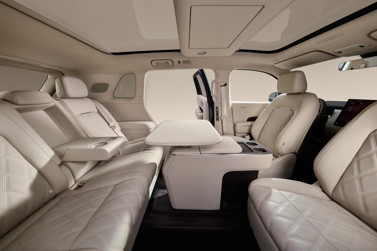 The luxury car interior features beige leather seats and a central armrest, which includes cup holders and a small table. The overall design is simple and modern. The interior space is spacious, with natural light coming in from the windows.