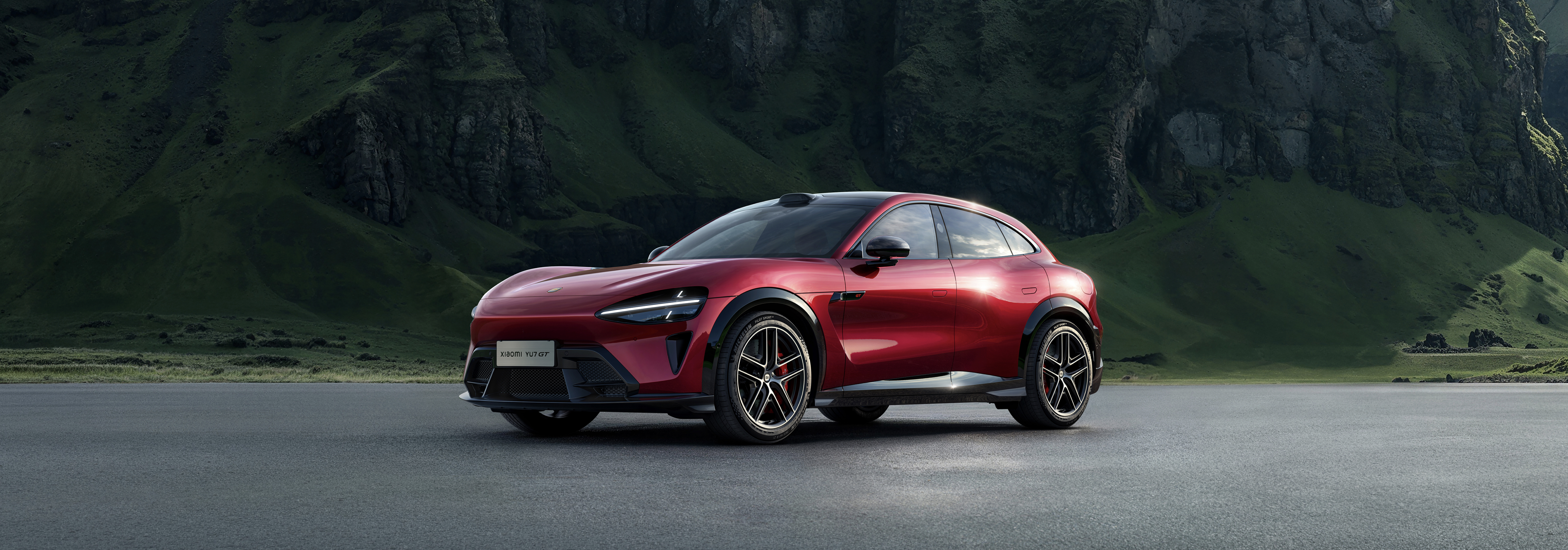 The red electric vehicle is parked on a flat road, with a background of green hills and rocks, showcasing the combination of natural scenery and modern technology.