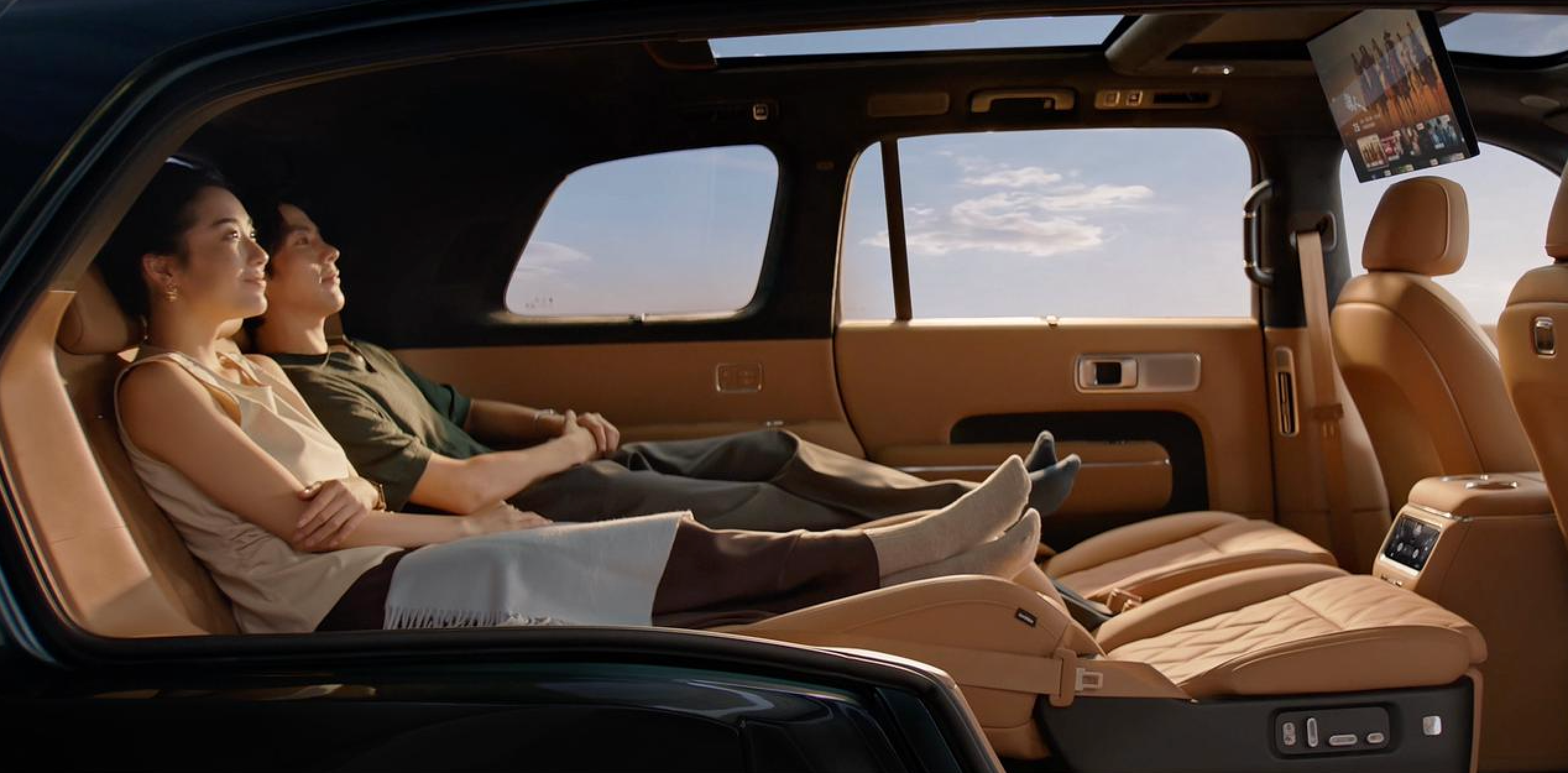 A man and a woman are sitting in a luxury car, enjoying a relaxing time, with a large screen display beside them and a blue sky in the background. Both are dressed casually, wearing socks, and the atmosphere is comfortable and pleasant.