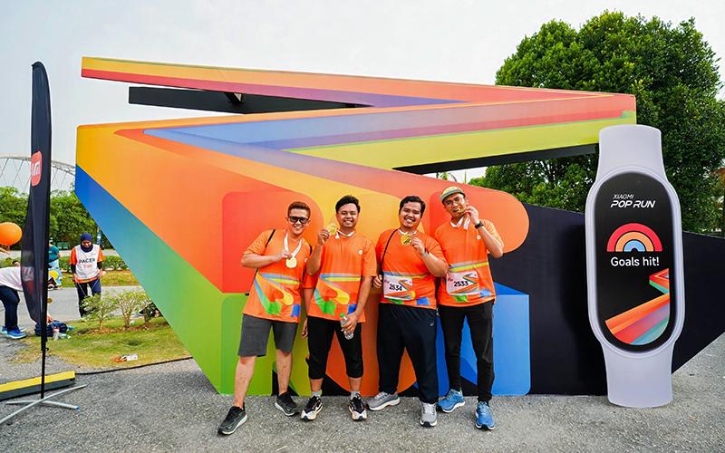 Several participants dressed in orange sportswear are posing for a photo in front of a colorful background, next to an electronic device displaying "Goals hit!" The setting is an outdoor activity venue, surrounded by green trees.