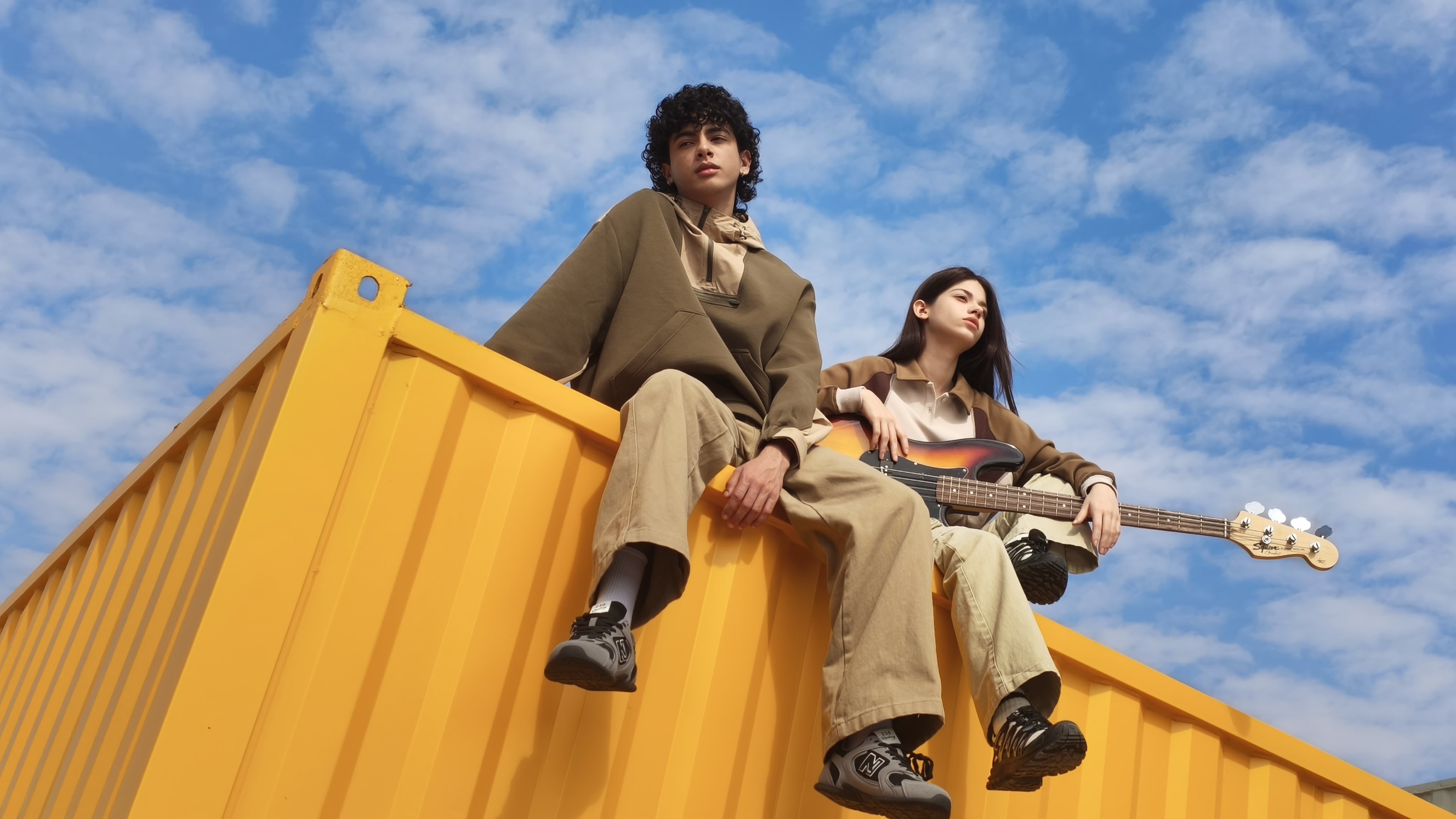 Two people are sitting on the edge of a yellow shipping container, one wearing a loose-fitting top, while the other holds an instrument, with a background of blue sky and white clouds.