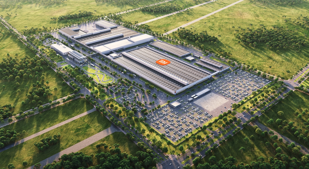 An aerial view of a modern factory, surrounded by vast green fields and trees. The factory building has a prominent sign, next to which is a large parking lot with vehicles parked neatly.