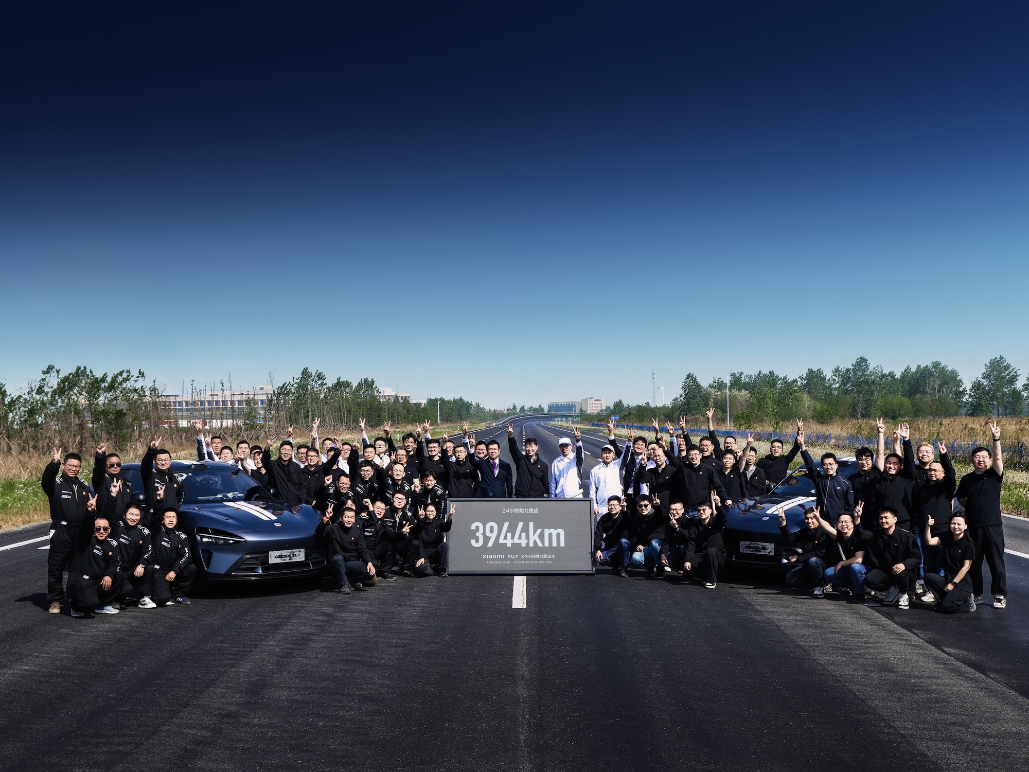 A group of people is standing on the road, dressed in black clothing, raising their hands in celebration, with two sports cars parked nearby. In front, there is a sign that reads 