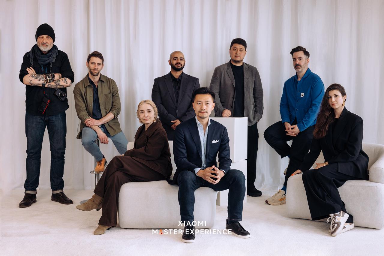 A group of people is posing for a photo in front of a white background, featuring several fashionable individuals, with a piece of white furniture nearby, creating a modern atmosphere.