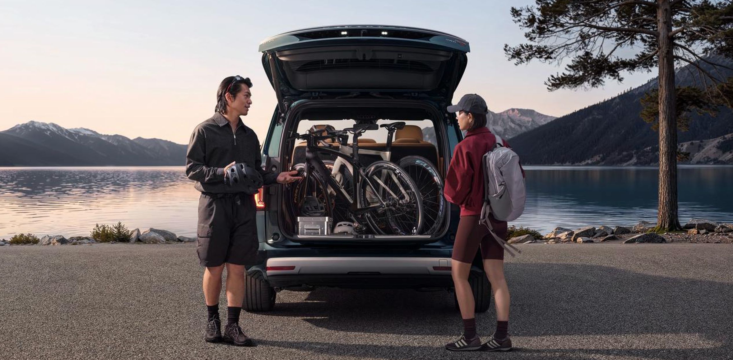 A man and a woman are standing next to a car with its trunk open, which is filled with several bicycles. The background features lakes and mountains, creating a peaceful and natural environment. The man is holding a helmet and seems to be talking to the woman.