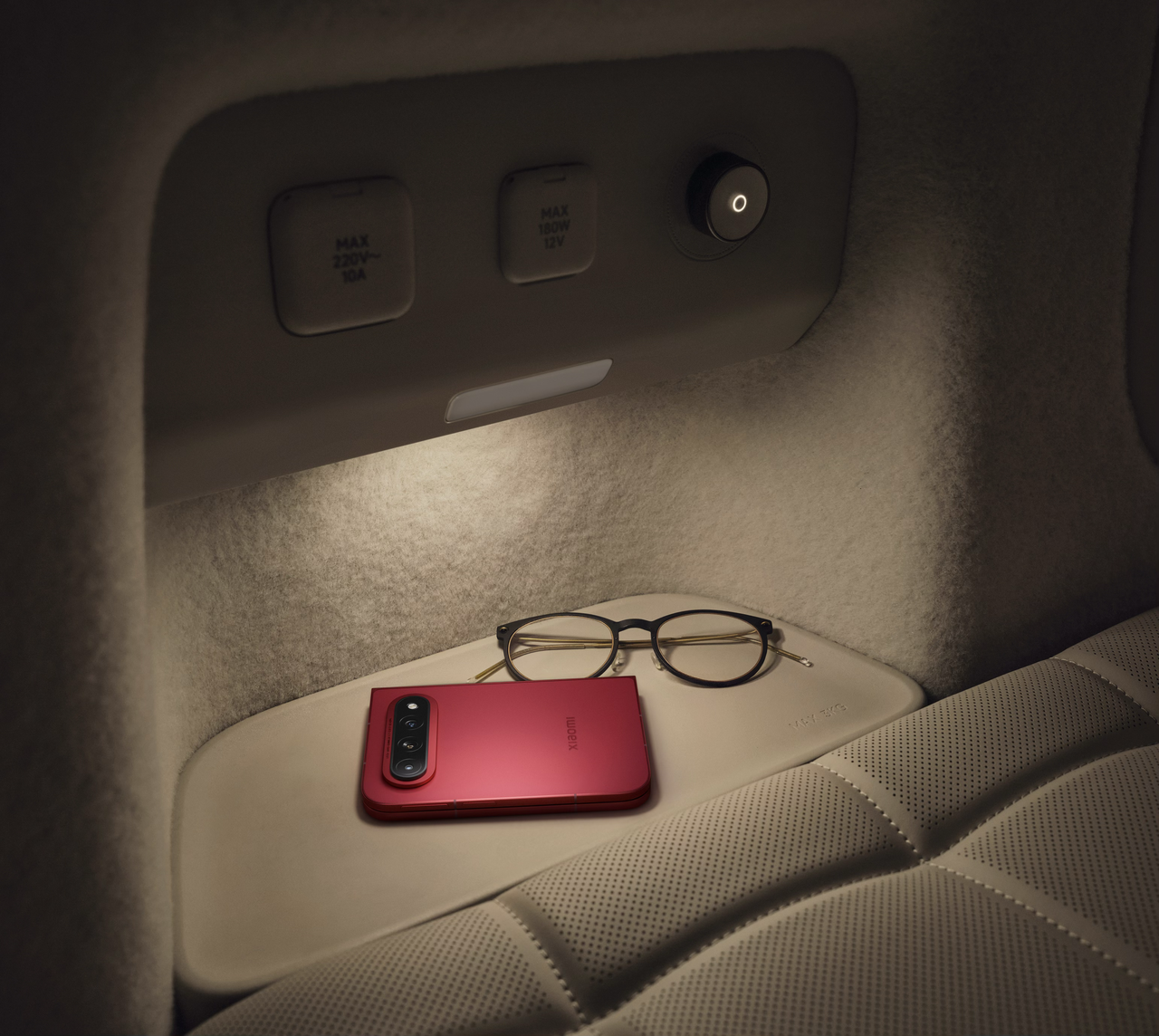 A red phone and glasses are placed on a soft surface, surrounded by lighting, with the background showing details of the car interior.