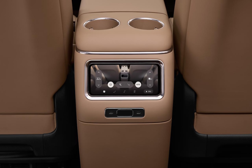 The rear central console of the car is equipped with a touchscreen display that shows the temperature settings and other function buttons. Surrounding it is beige leather upholstery, with two cup holders in the center.