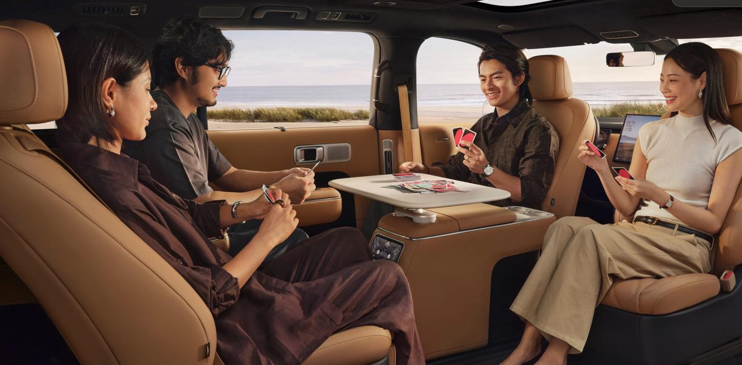 Four people are sitting in the back seat of a luxury vehicle, playing cards around a foldable table. The interior of the car is beige, and outside the window, there is a view of the beach and the ocean.