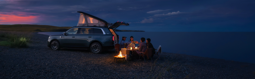 A group of people is sitting around a campfire on the beach, enjoying their camping time. Next to them is an SUV with its tailgate open, surrounded by the tranquility of the night and the beautiful sky.